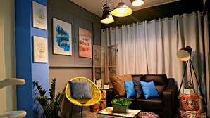 A modern living room with a black leather sofa, yellow chair, and various decorative items including a lamp, potted plants, and framed artwork. - Transit Point Hostel Mactan Cebu (Lapu-Lapu)
