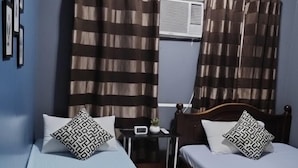 A room with two single beds, each with patterned bedding and pillows, a small bedside table with a clock, and a window with curtains. - Transit Point Hostel Mactan Cebu (Lapu-Lapu)