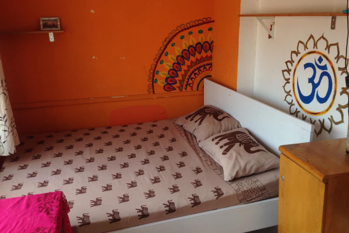 A small, single-bed room with a patterned bedspread, a headboard with an 'Om' symbol, and a wall with a decorative design.
