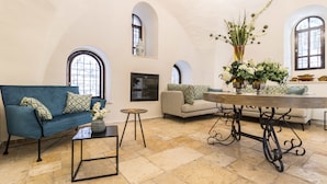 Un salón espacioso con un sofá azul, una mesita redonda y una mesa de comedor con un jarrón de flores. Hay dos ventanas con arcos y una chimenea.