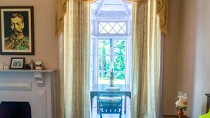A room with a large window, a fireplace, a table with a chair, and a portrait on the wall.