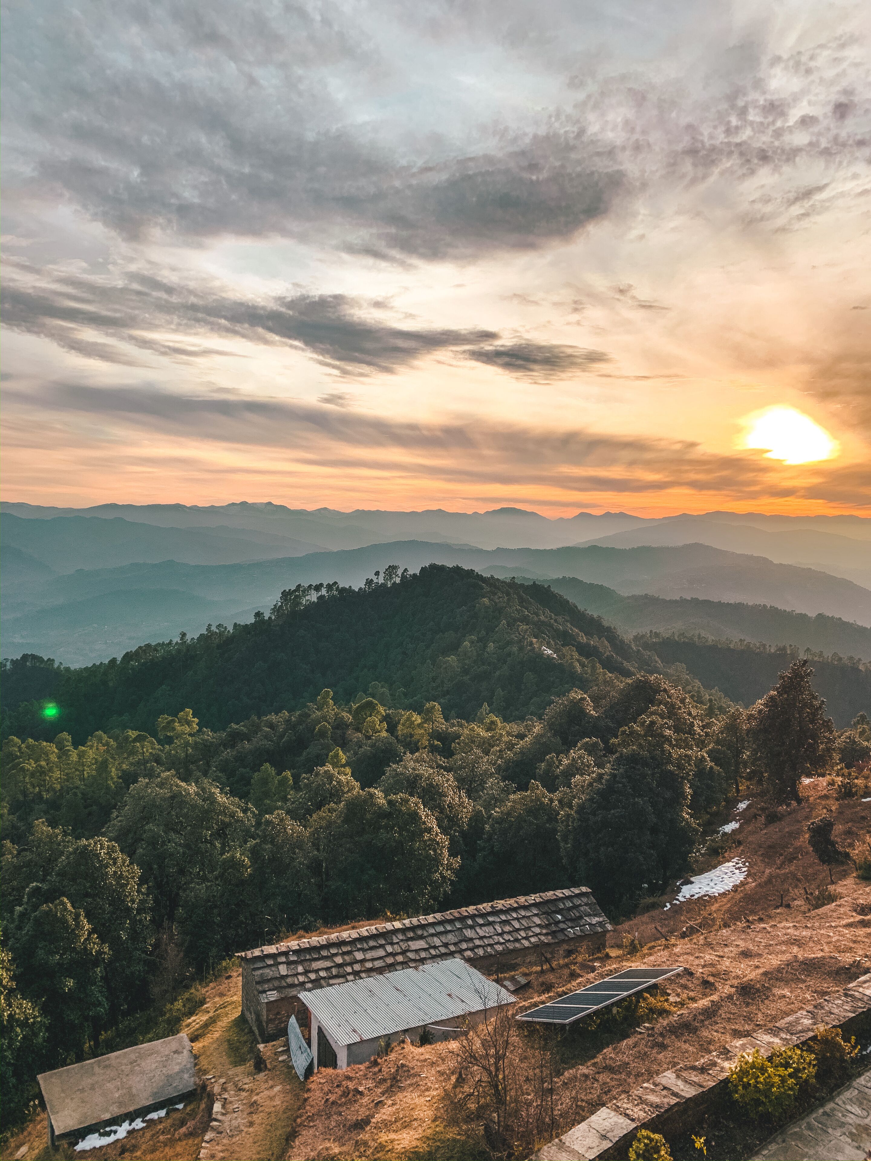 A rural landscape with a small building featuring a sloped roof and solar panels, surrounded by dense forest and distant mountains under a cloudy sky.