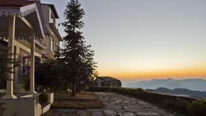 A stone pathway leading to a building with a balcony, surrounded by trees and mountains in the background.