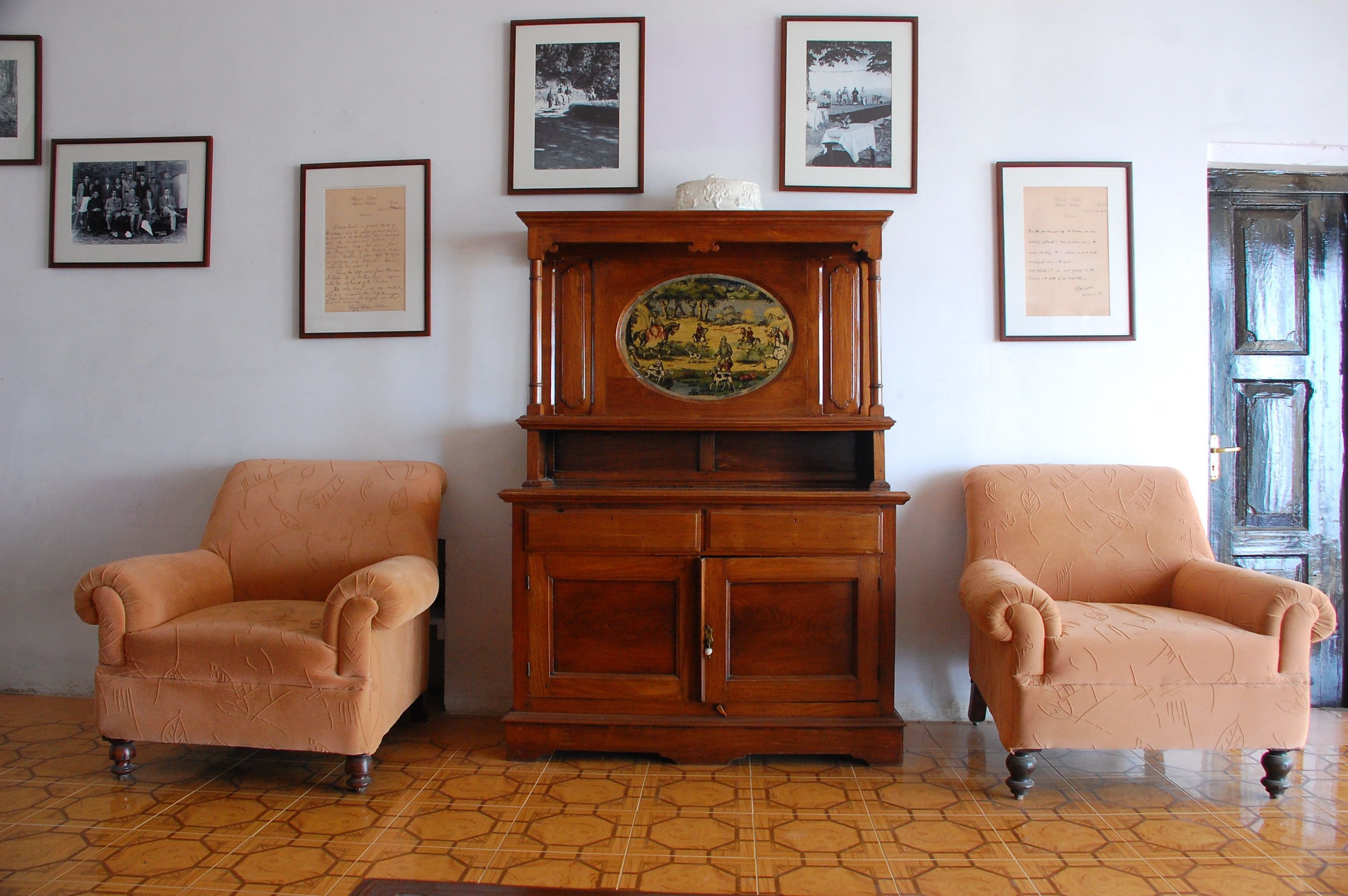 A room with two armchairs, a wooden cabinet with a decorative plate, and framed documents on the wall.