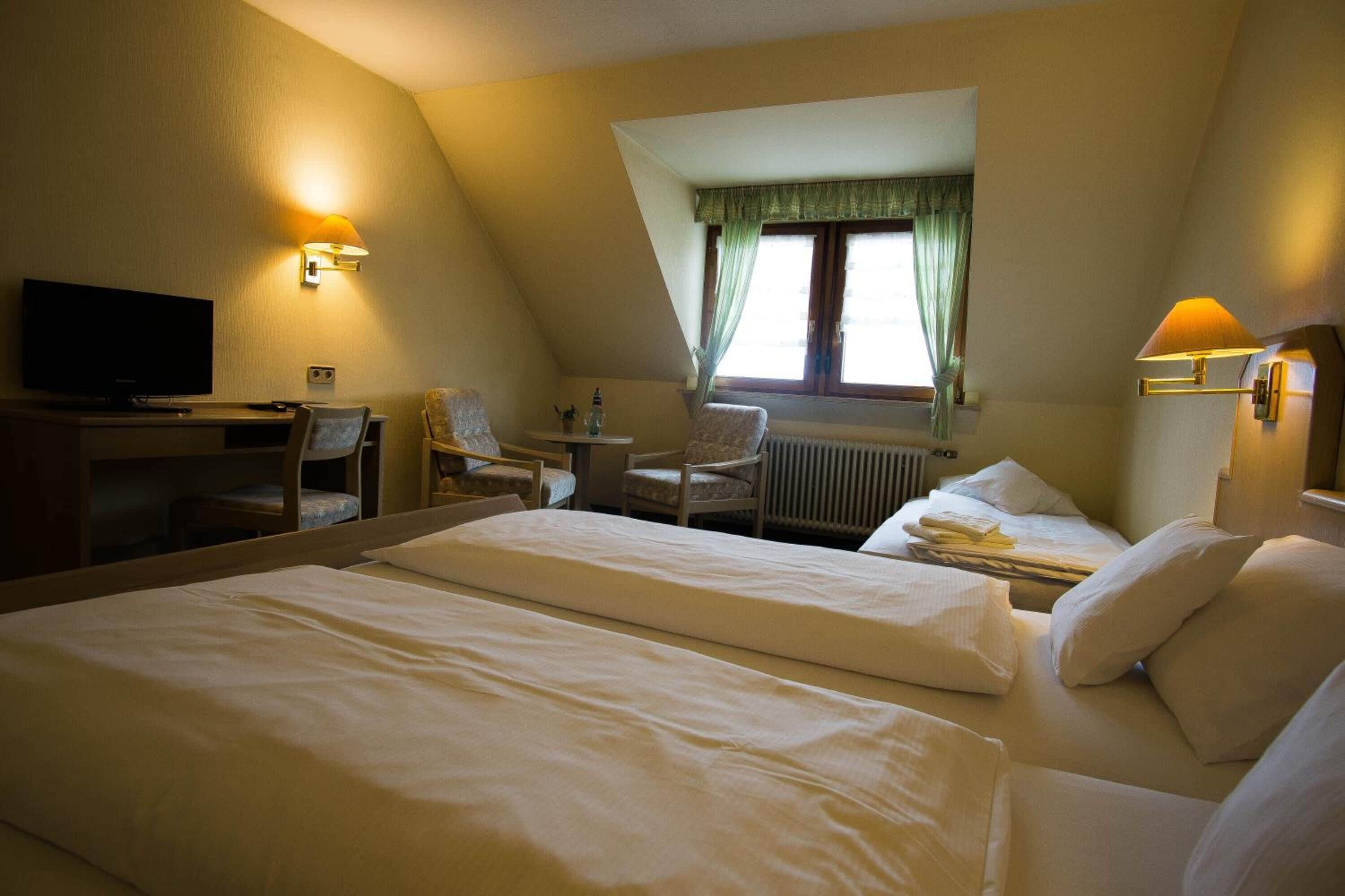 Habitación de hotel con dos camas, un escritorio con televisor, una silla y una ventana con cortinas.