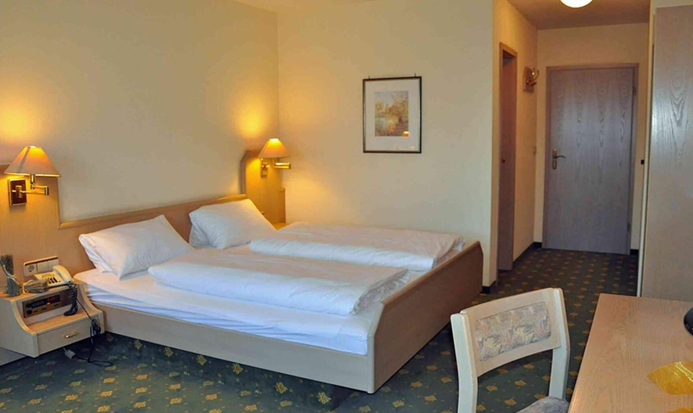 A hotel room with a double bed, bedside lamps, a telephone, and a small table with a chair.