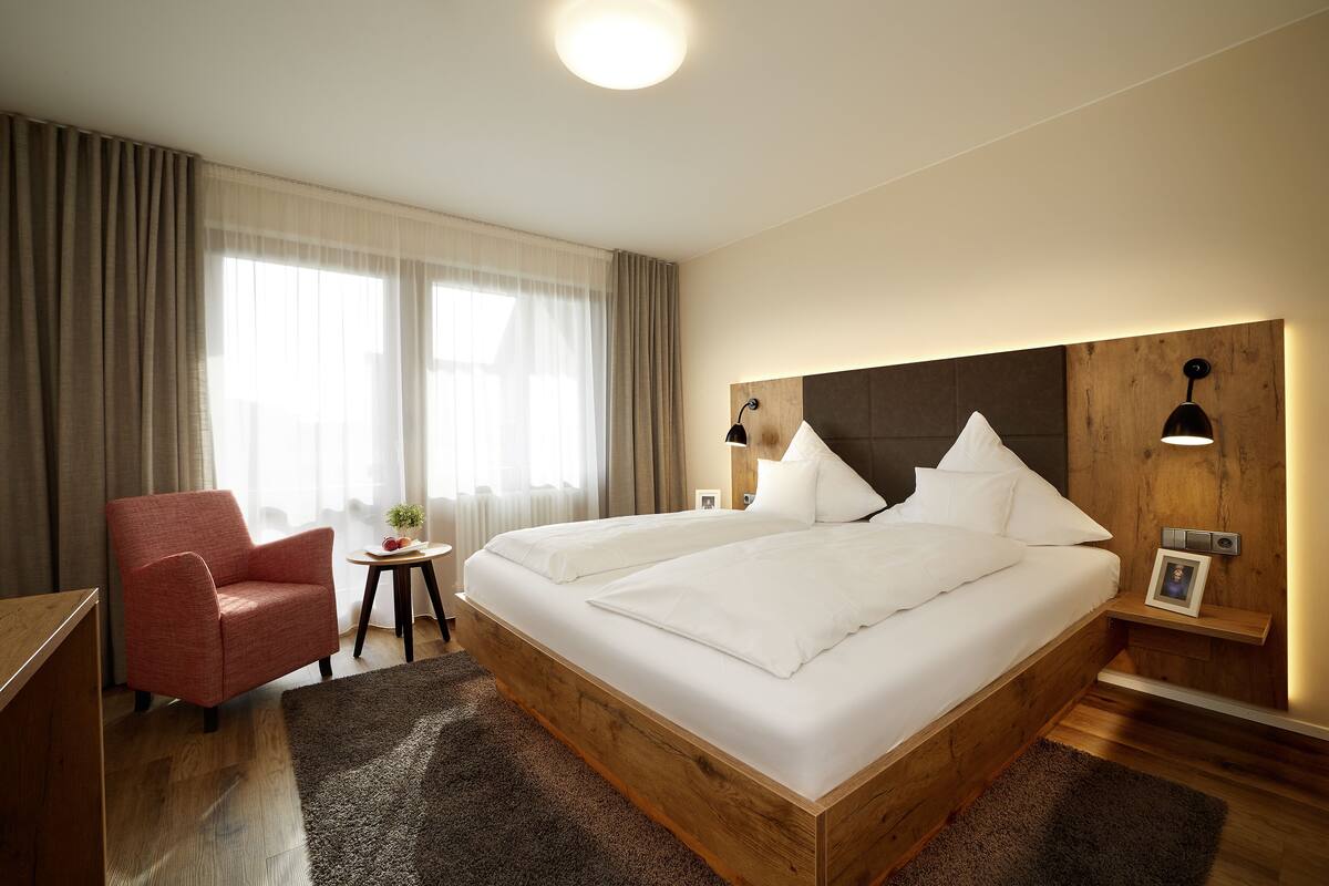 Una habitación de hotel moderna con una cama grande, un sillón rojo, una mesita con una planta y una lámpara de noche.