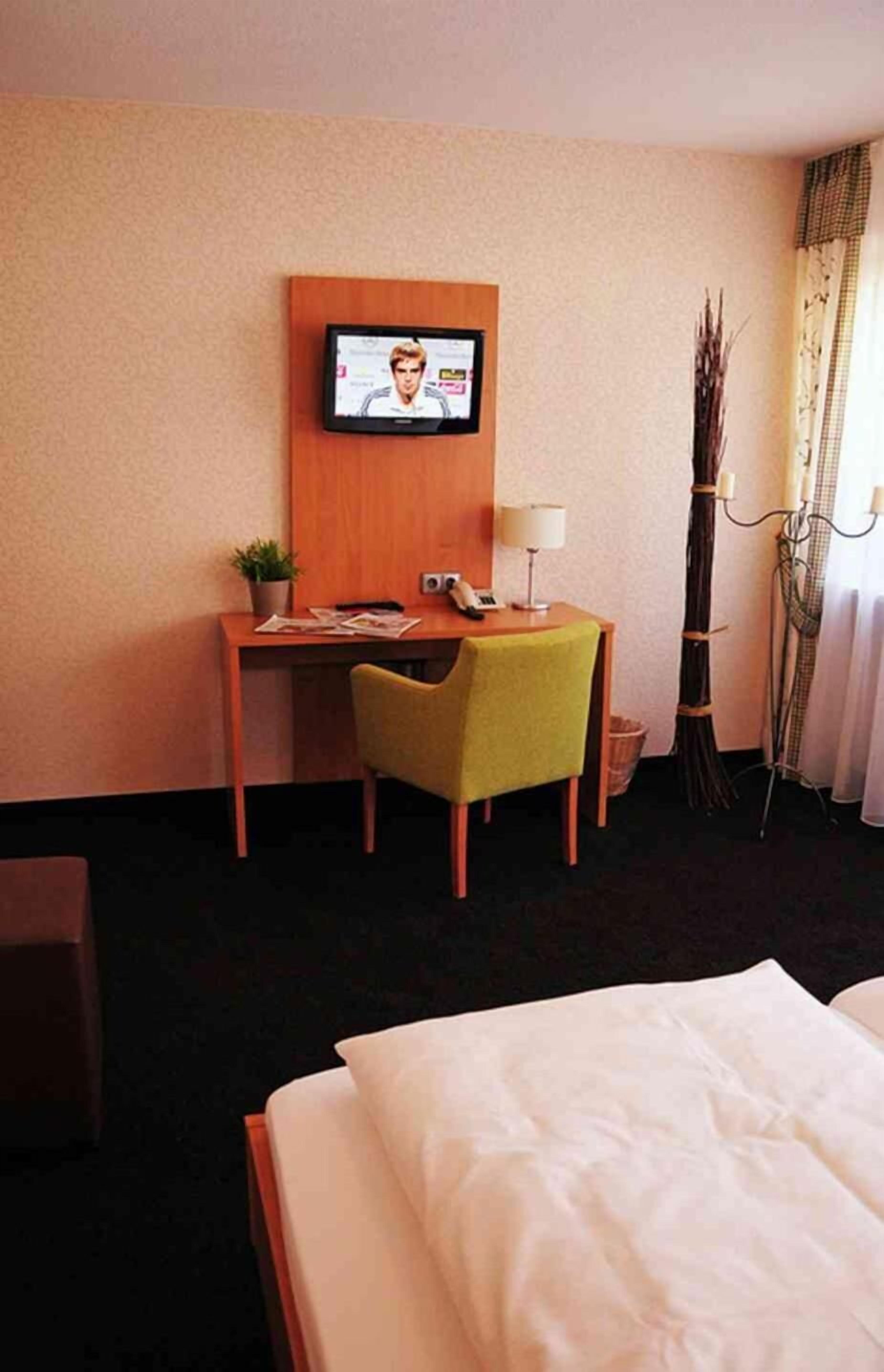 Ein Hotelzimmer mit einem Bett, einem Schreibtisch mit Stuhl, einem an der Wand befestigten Fernseher, einer Lampe und einer Zimmerpflanze.