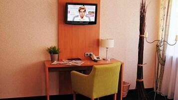 Ein Hotelzimmer mit einem Bett, einem Schreibtisch mit Stuhl, einem an der Wand befestigten Fernseher, einer Lampe und einer Zimmerpflanze.