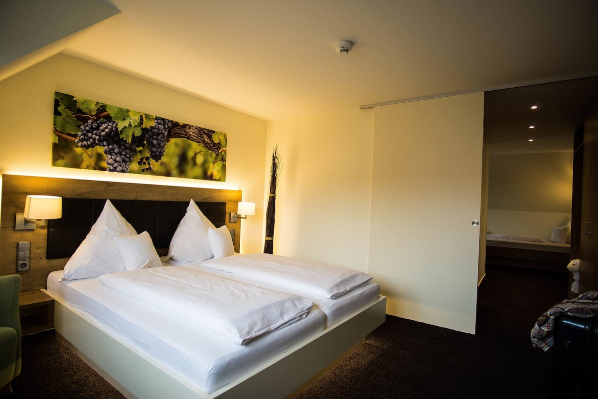 Una habitación de hotel con una cama grande, un cabecero con una pintura de parra, una mesita de noche con lámpara y una puerta que da a otra habitación.