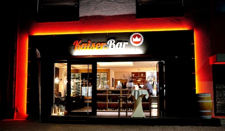 Un escaparate con un letrero que dice "Kaiser Bar" y un logotipo con una corona. En el interior, hay estantes con diversos artículos y una zona de mostrador.