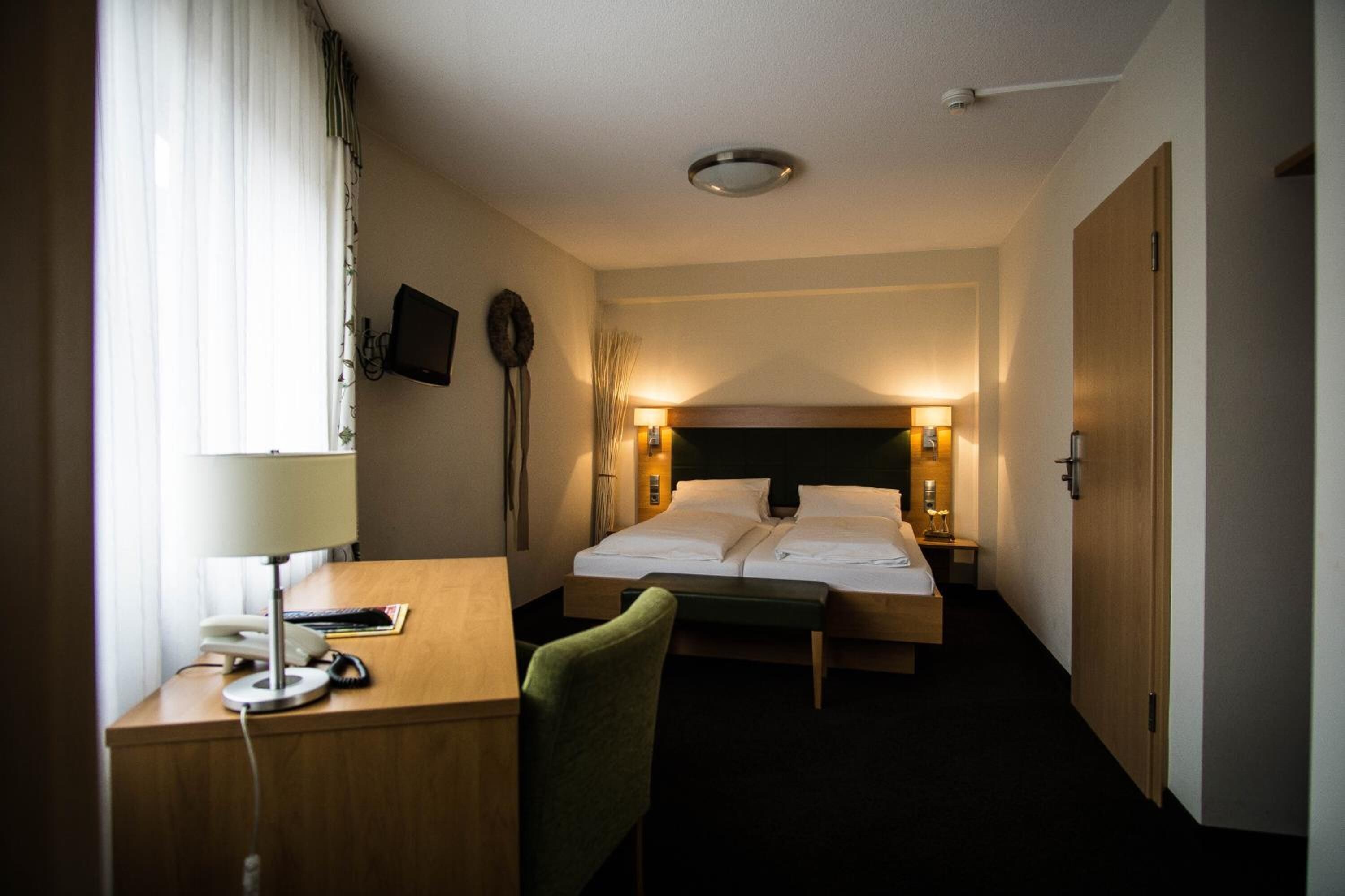 Habitación de hotel con cama, mesitas de noche, escritorio con lámpara, una silla y televisión en la pared.