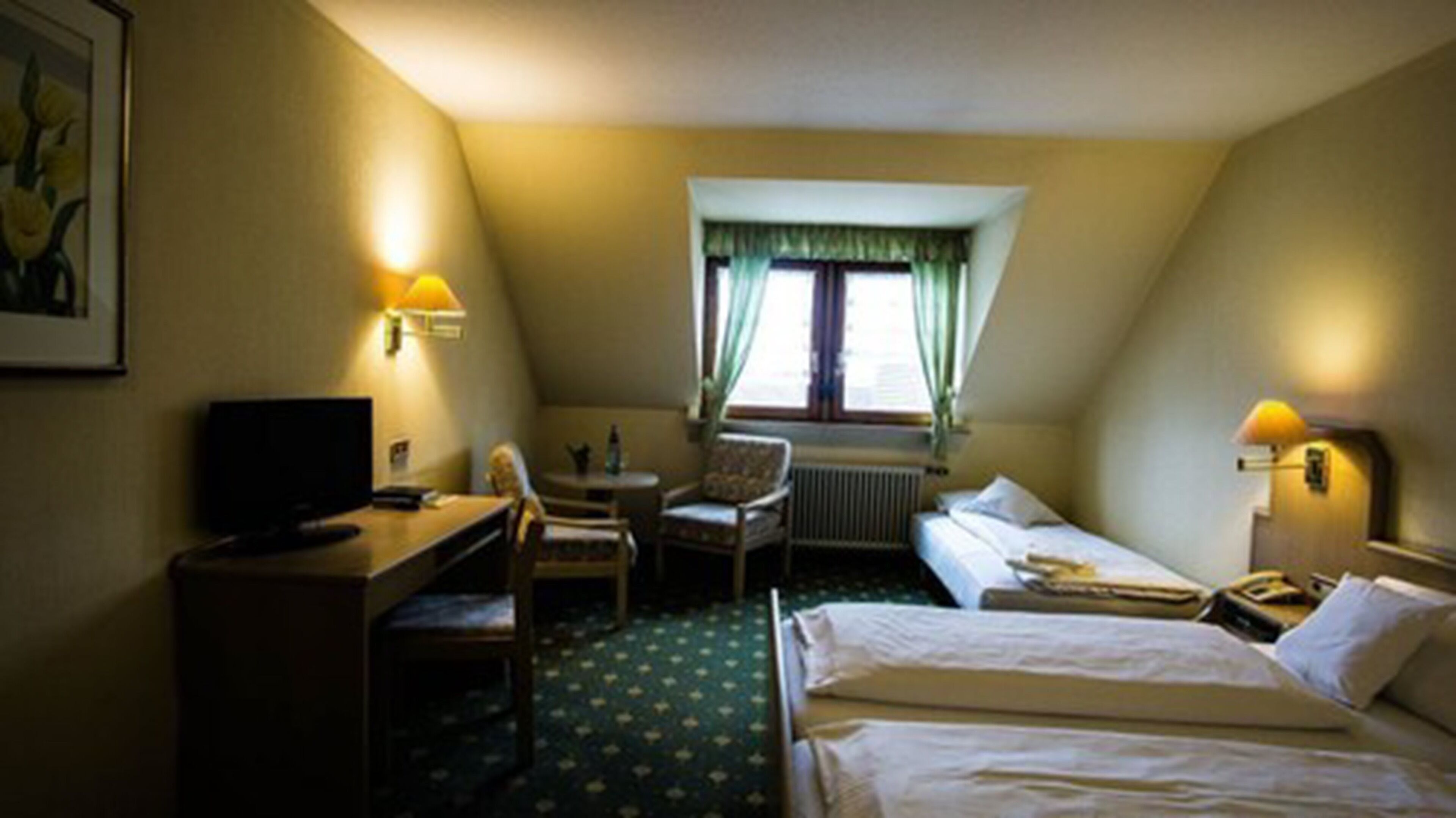 Une chambre d’hôtel équipée d’un bureau, d’une chaise, de deux lits, d’un radiateur et d’une fenêtre avec des rideaux.