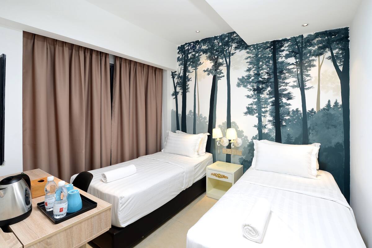 Une chambre d’hôtel avec deux lits, un bureau doté d’une bouilloire et de bouteilles d’eau, et un tableau représentant des arbres accroché au mur.
