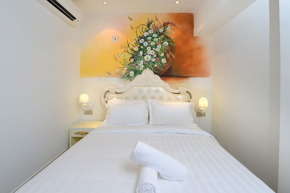 Un lit bien fait, avec une serviette blanche roulée, une tête de lit décorative et un tableau représentant des fleurs accroché au mur.