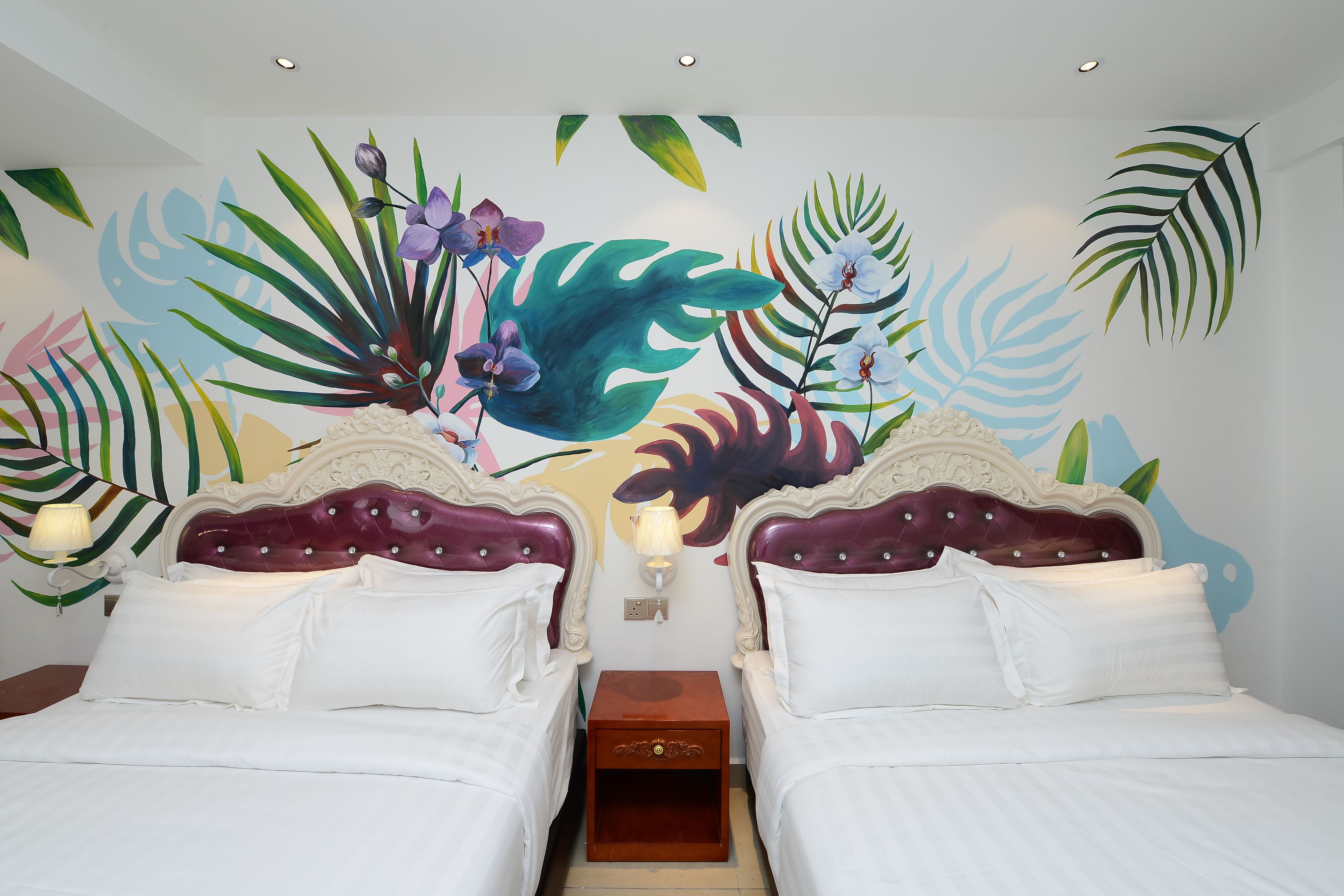 A hotel room with two beds, white linens, a wooden nightstand, a wall mural with tropical leaves and flowers, and a ceiling light.