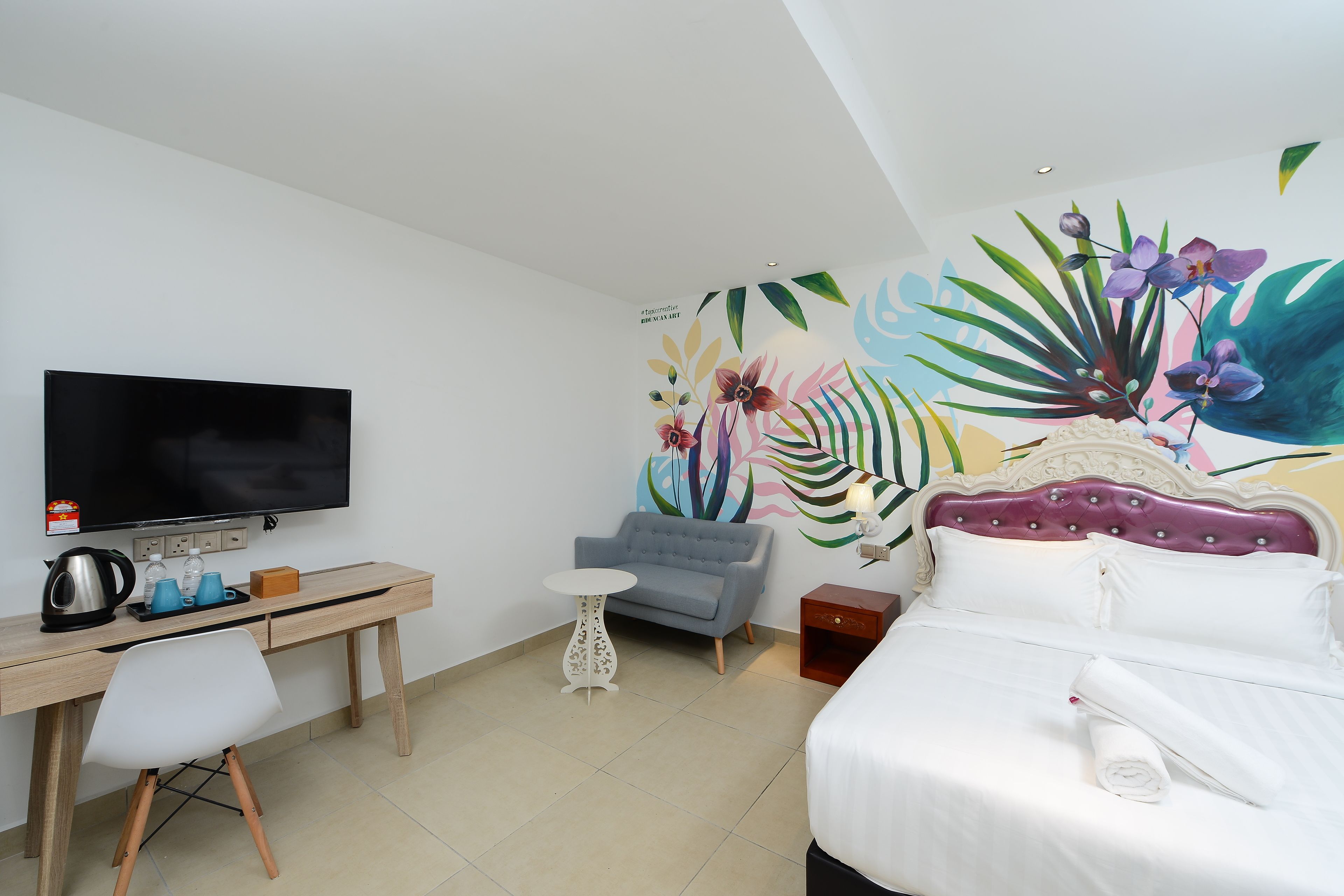 Habitación de hotel con cama, escritorio con televisor, silla, mesita y un mural de pared colorido.