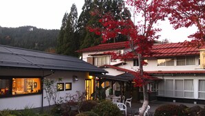 A two-story building with a red roof, a covered patio area with white chairs, and a garden with various plants and a tree with red leaves.