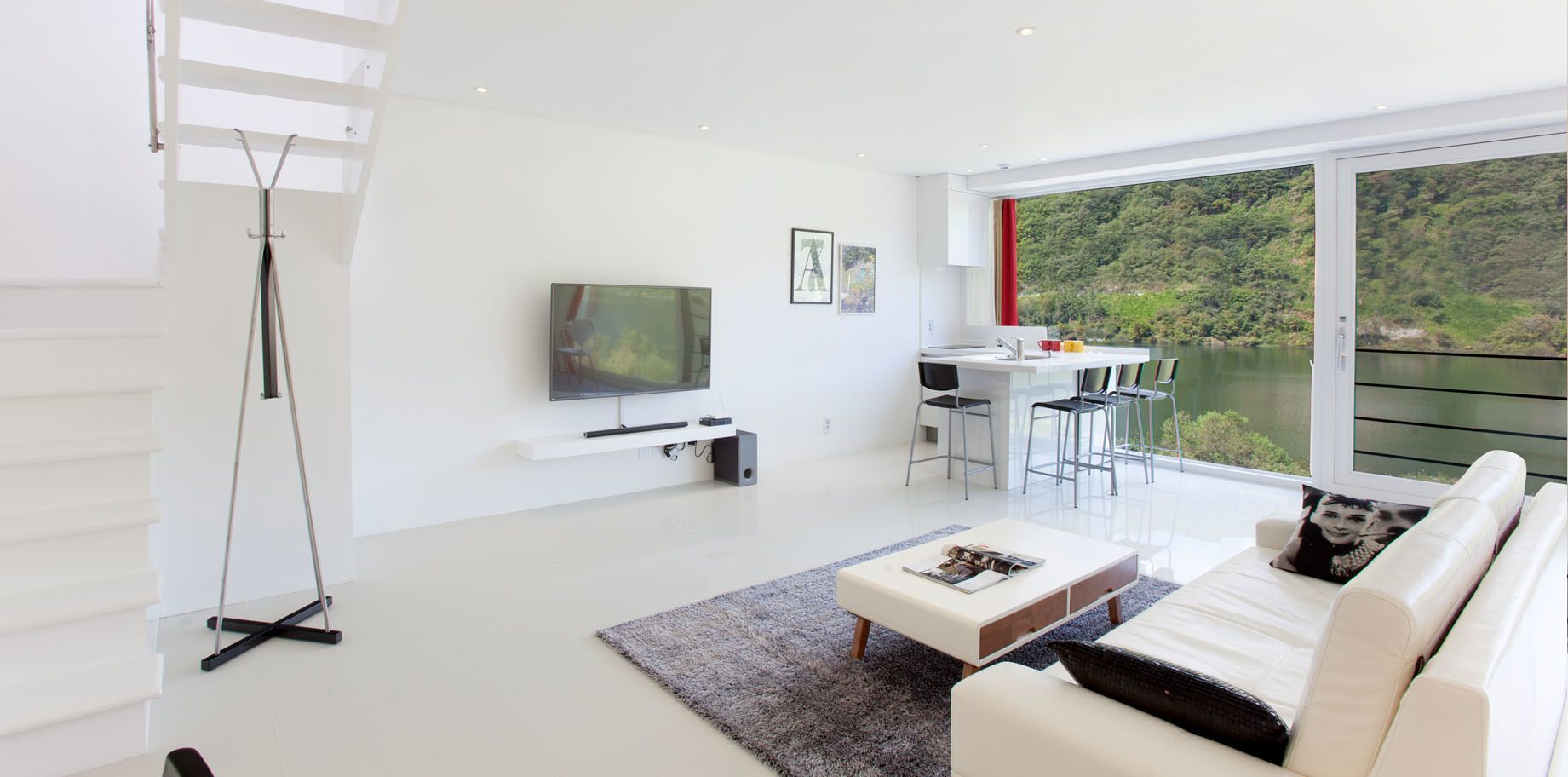 A modern living room with a flat-screen TV, a dining area with a table and chairs, and a large window offering a view of a lake and greenery.