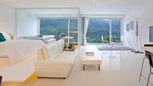 Une chambre d’hôtel moderne avec un grand lit, un canapé blanc, une petite table et une vue sur un paysage montagneux grâce à de grandes fenêtres.