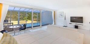 A modern hotel room with a large bed, a TV, a balcony with a pool view, and a dining area.