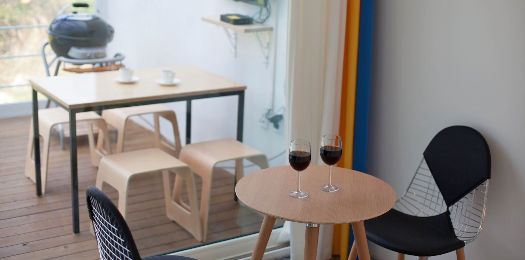 A modern outdoor dining area with a round table and two chairs, two glasses of red wine, and a barbecue grill in the background.