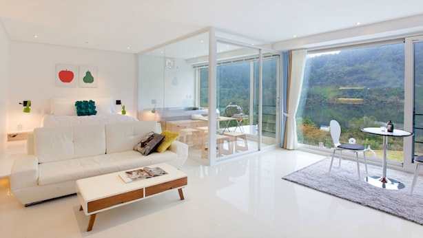 Un salon moderne avec un canapé blanc, une table basse et un coin repas comprenant une table et des chaises. De grandes fenêtres offrent une vue sur un paysage boisé.