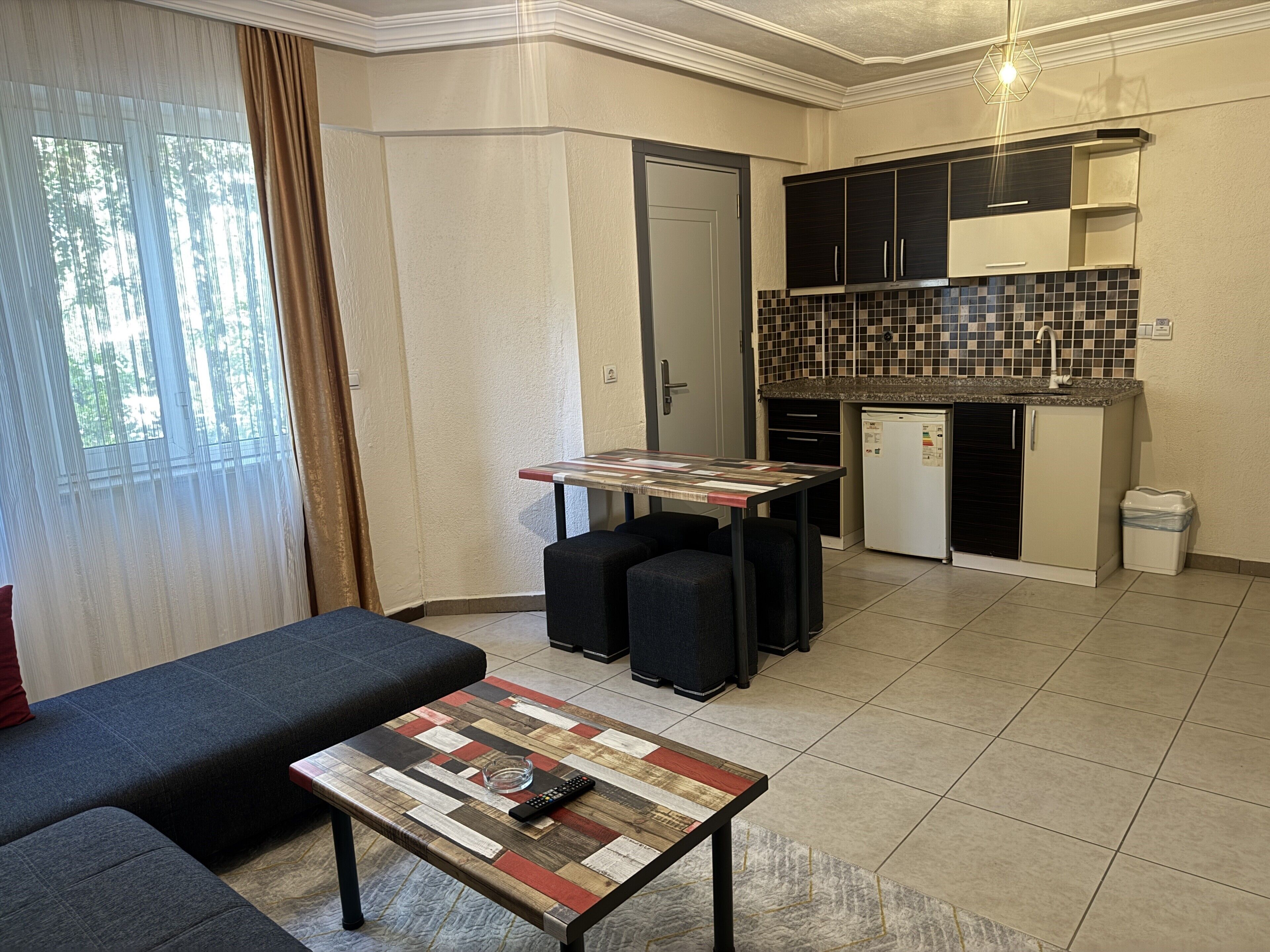A compact living space with a sofa, a coffee table, a small kitchen area with a tiled backsplash, and a door leading to another room.