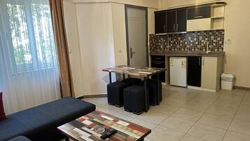 A compact living space with a sofa, a coffee table, a small kitchen area with a tiled backsplash, and a door leading to another room.