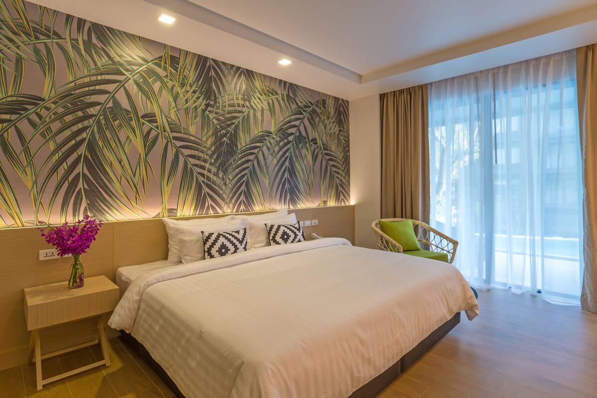 A modern hotel room with a large bed, a nightstand with a vase of flowers, a wall with a tropical leaf pattern, a green chair, and a large window with curtains.