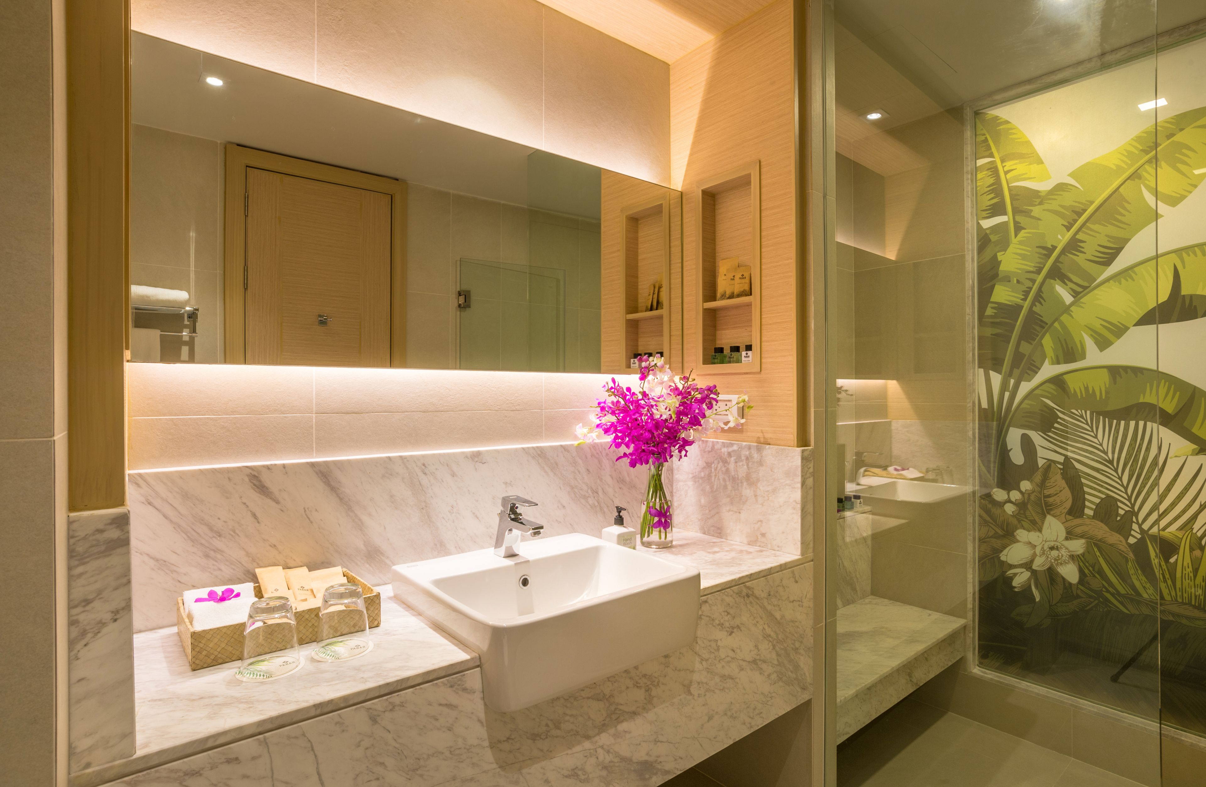 A modern bathroom with a large mirror, two sinks, marble countertops, and a shower with a tropical leaf design.