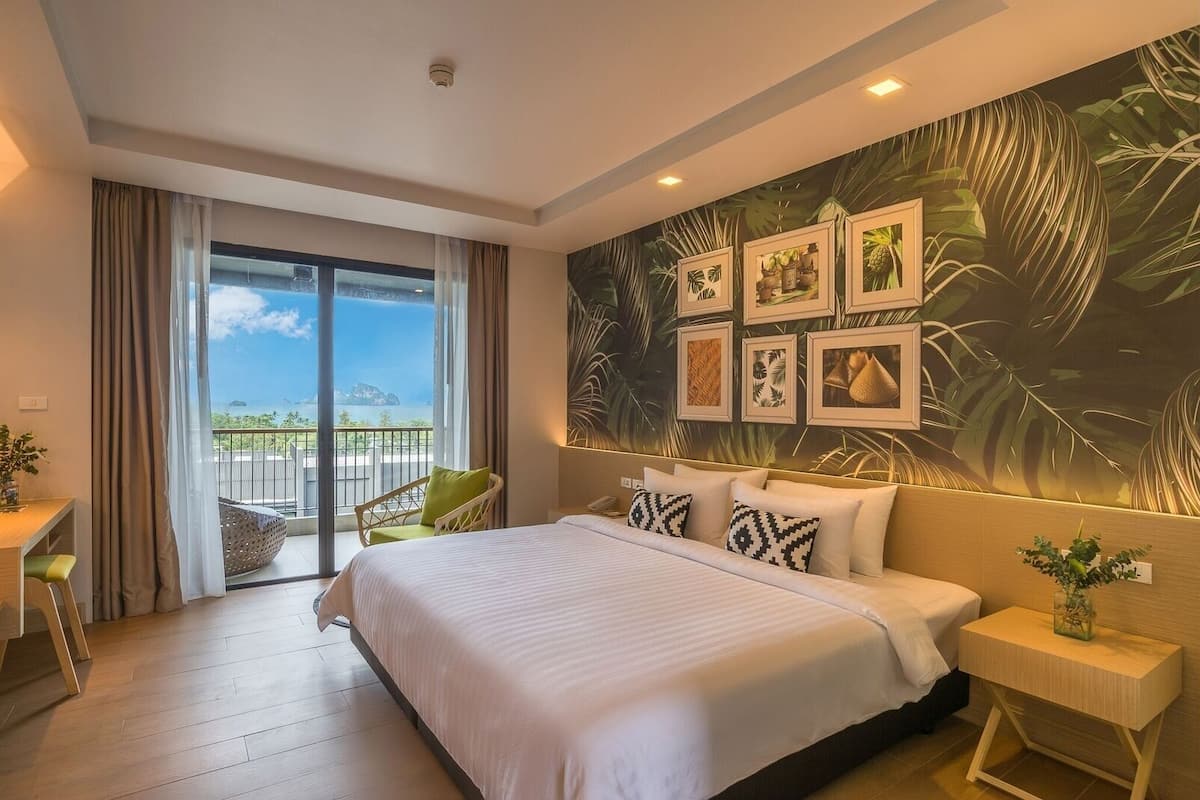 A hotel room with a large bed, a view of the sea through a sliding door, and tropical-themed wall art.