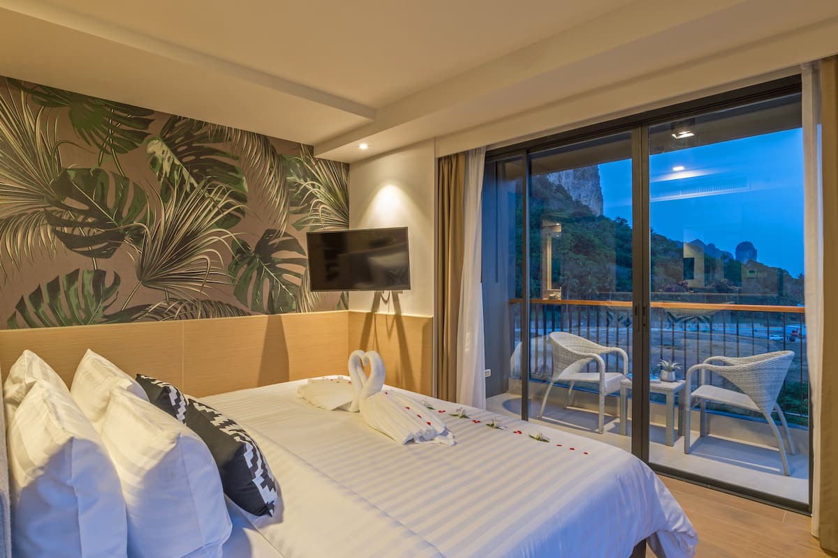 A hotel room with a bed, a flat-screen TV, a balcony with wicker chairs, and a scenic view.