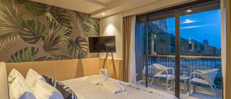 Habitación de hotel con cama, televisor de pantalla plana, balcón con sillas de mimbre y vistas panorámicas.