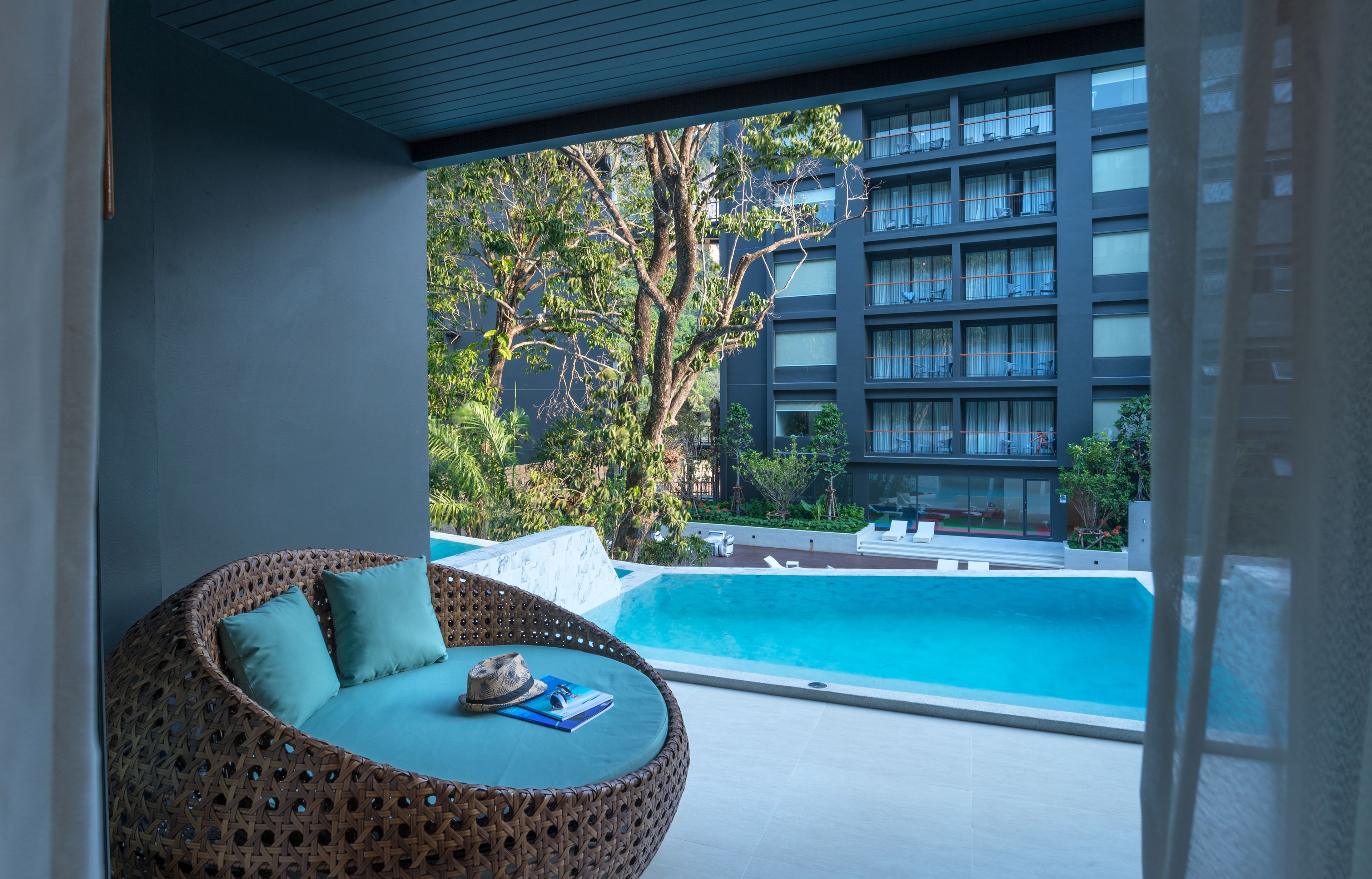 Una silla de mimbre con cojines y un sombrero sobre una mesa cerca de una piscina, con vista a un edificio con varios balcones.