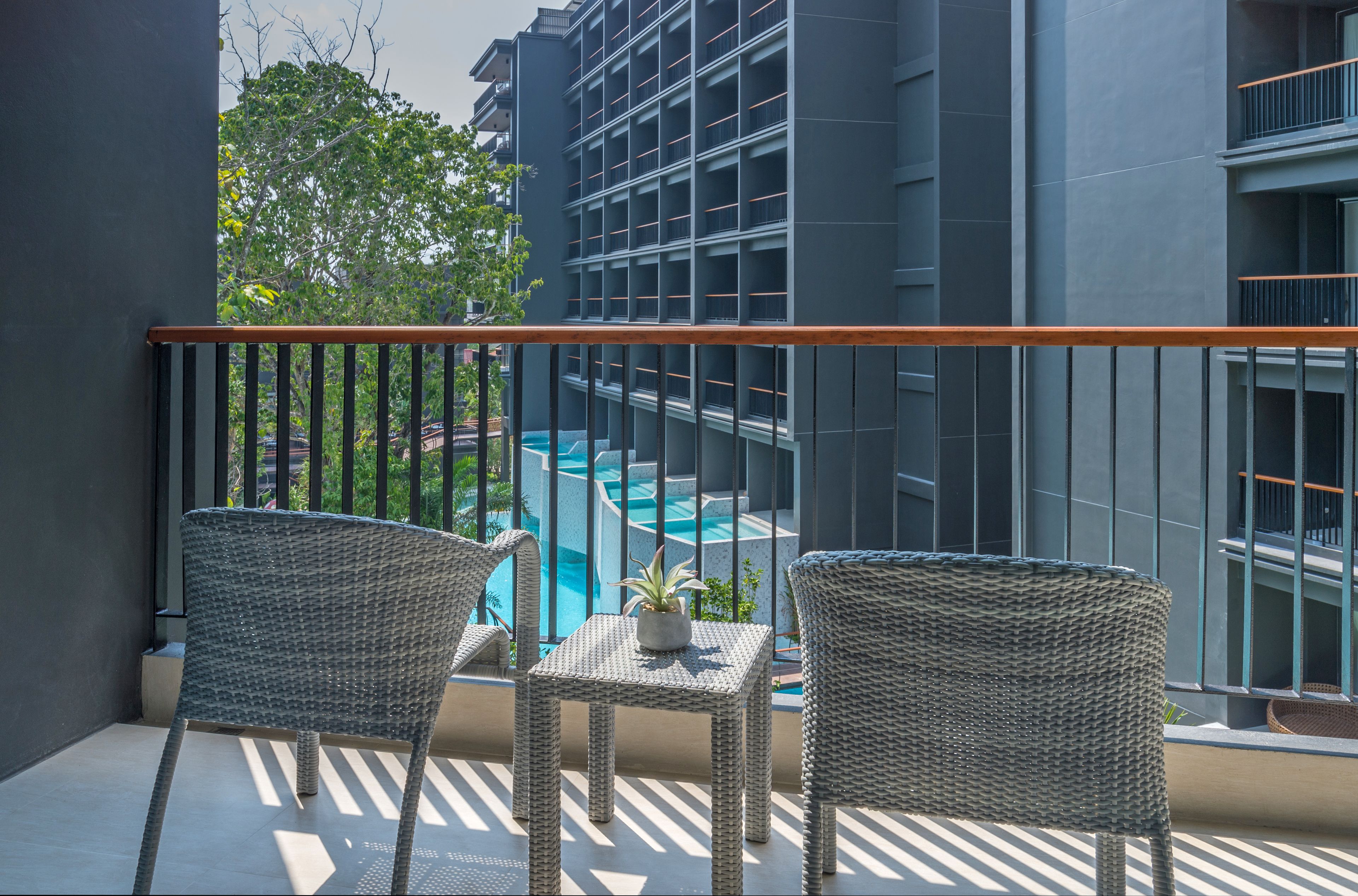 A balcony with wicker chairs and a small table, overlooking a swimming pool and a building with balconies.