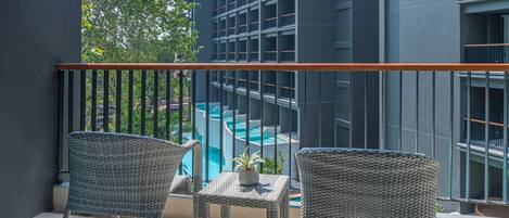A balcony with wicker chairs and a small table, overlooking a swimming pool and a building with balconies.