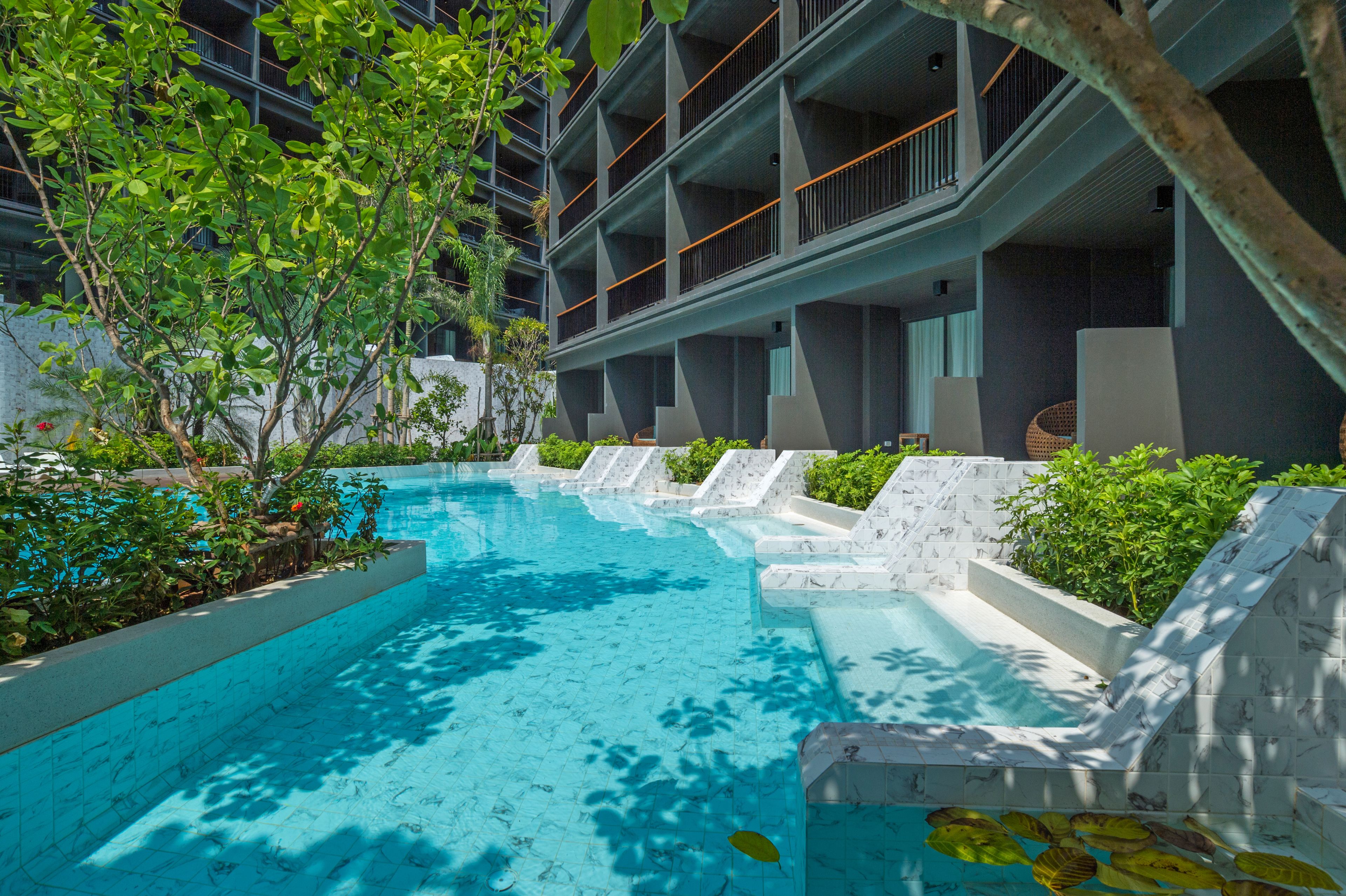 A swimming pool with a modern design, surrounded by greenery and a multi-story building with balconies.