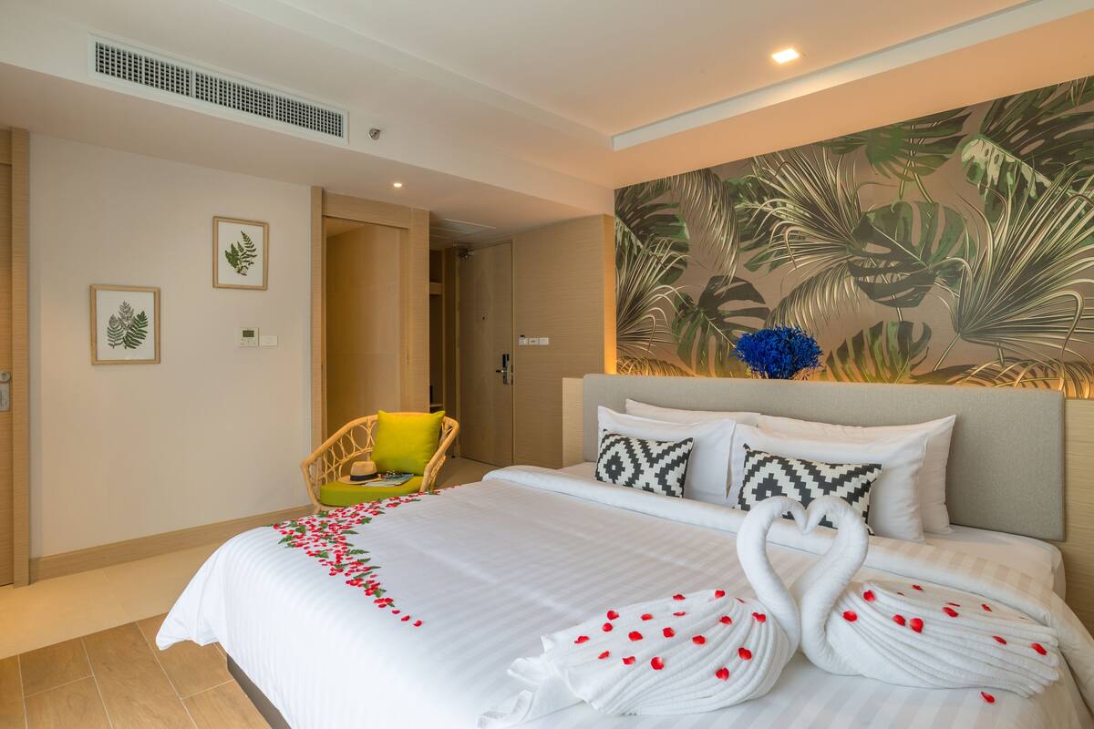 A hotel room with a large bed, a tropical leaf mural, a bright yellow chair, and a wall-mounted thermostat.