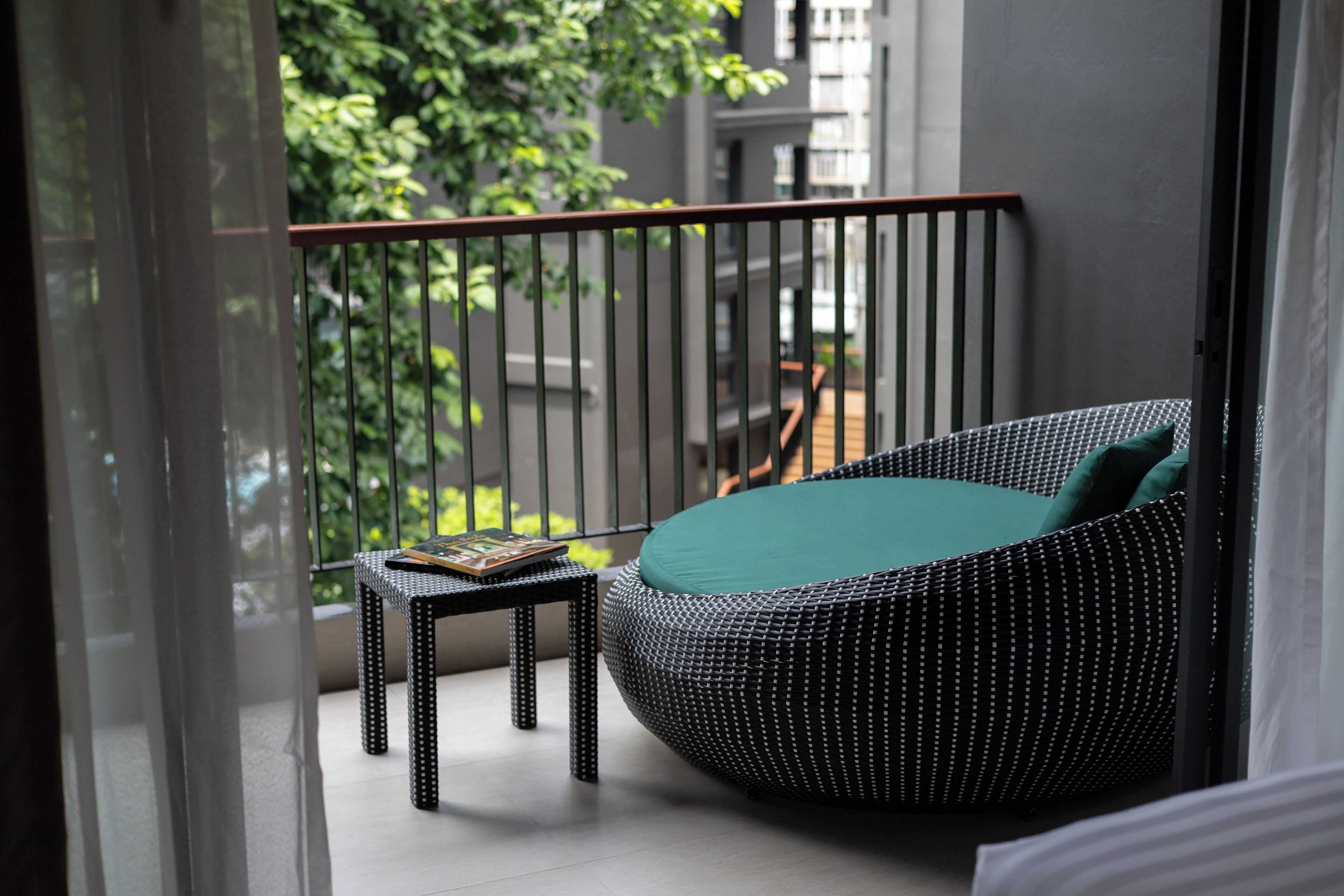 A balcony with a wicker sofa and a small table, overlooking a green garden and buildings.