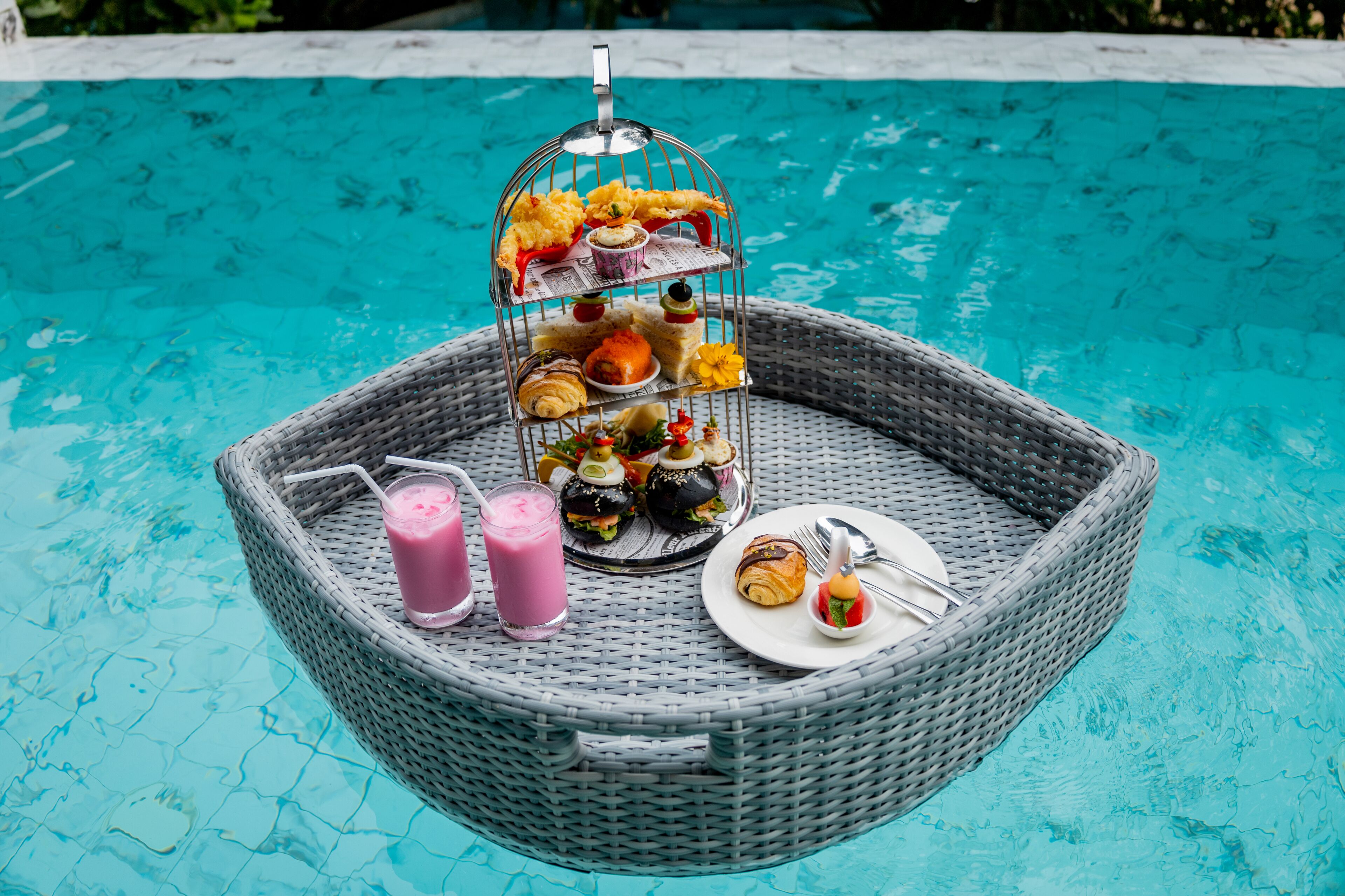 Une table en osier, avec de la nourriture et des boissons, placée au bord d’une piscine.