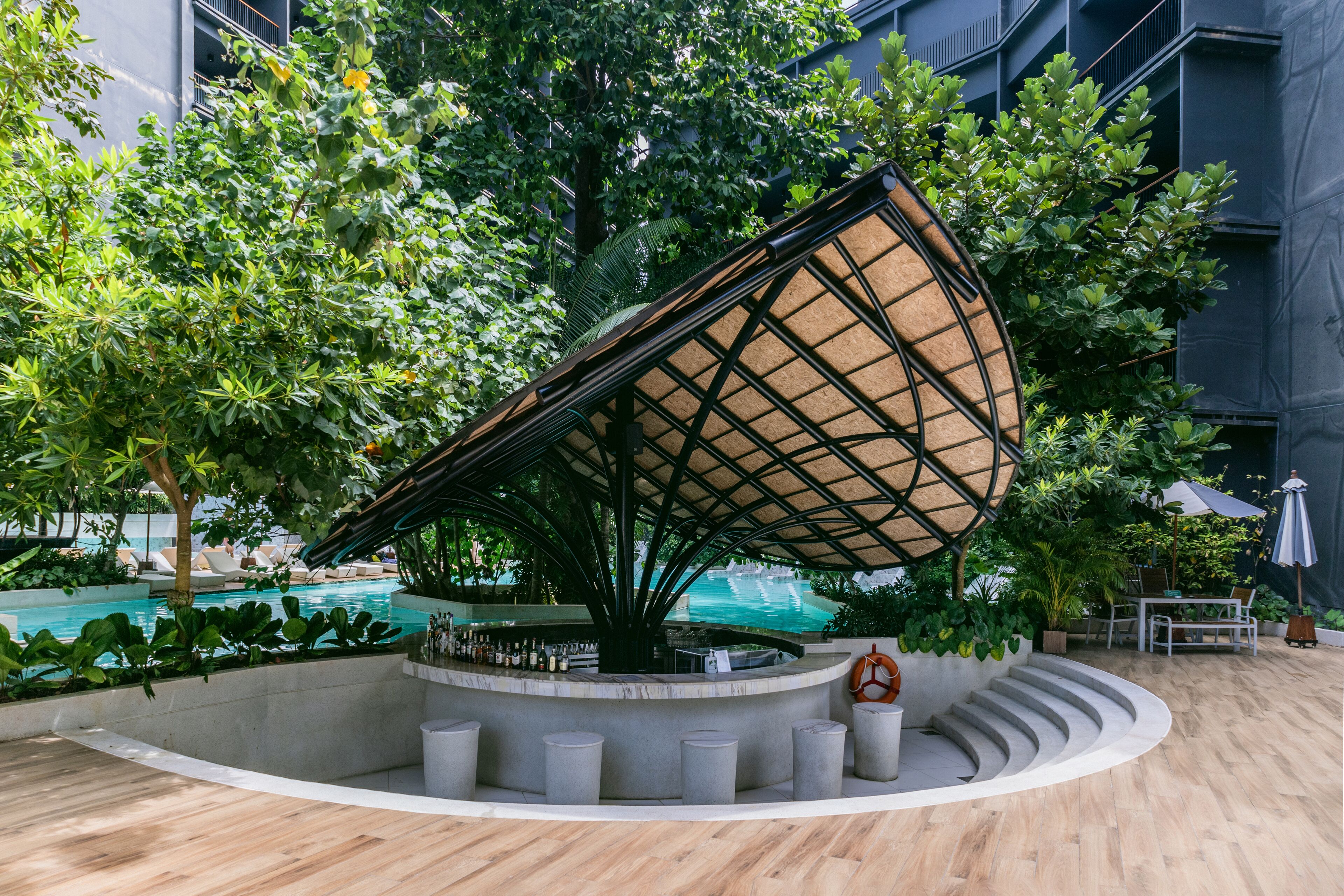 Zona de bar circular con un árbol en el centro, rodeada por una piscina y sillones de descanso.