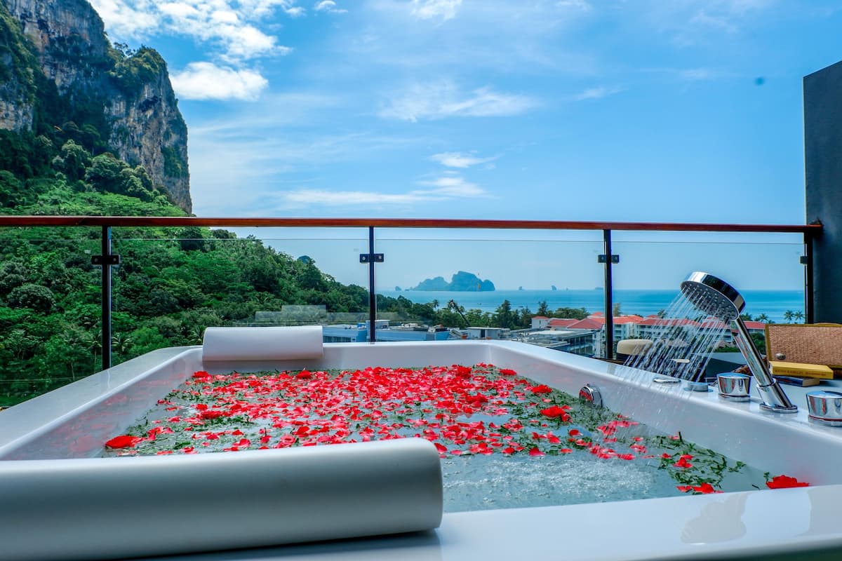 A hot tub with red rose petals, a showerhead, and a view of a mountainous coastline.