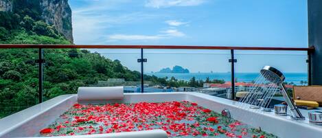 A hot tub with red rose petals, a showerhead, and a view of a mountainous coastline.
