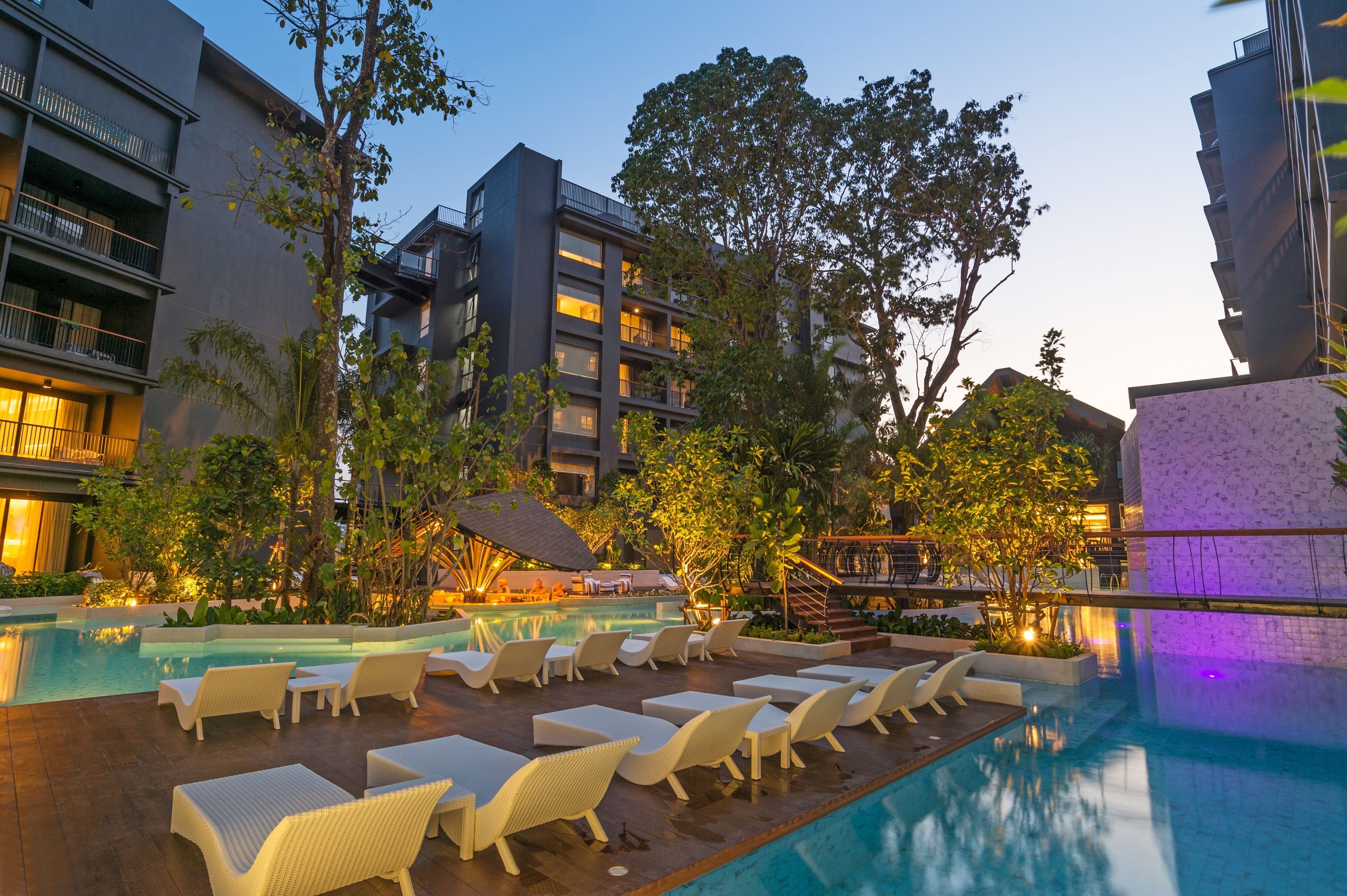 A hotel pool area with lounge chairs and a swimming pool surrounded by trees and buildings.
