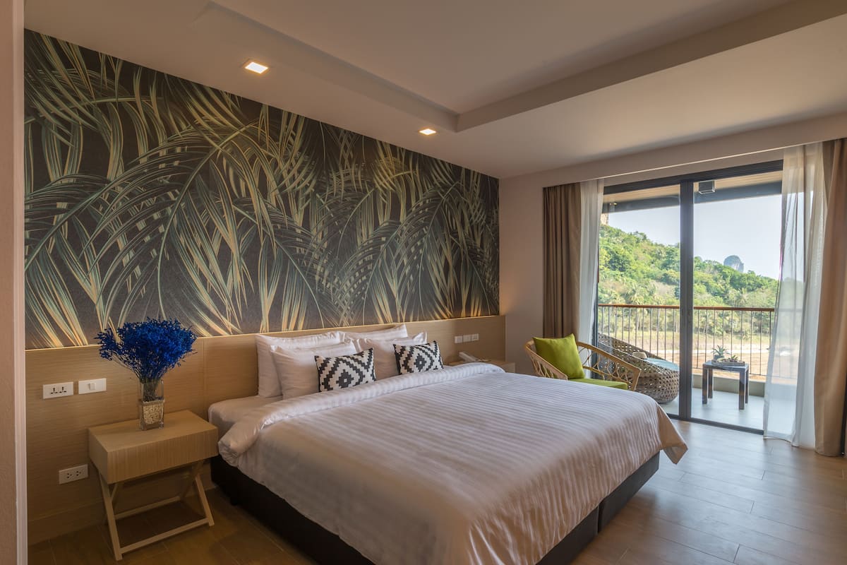 A spacious bedroom with a large bed, a nightstand, a vase with flowers, a view of greenery through a balcony door, and a tropical leaf pattern on the wall.