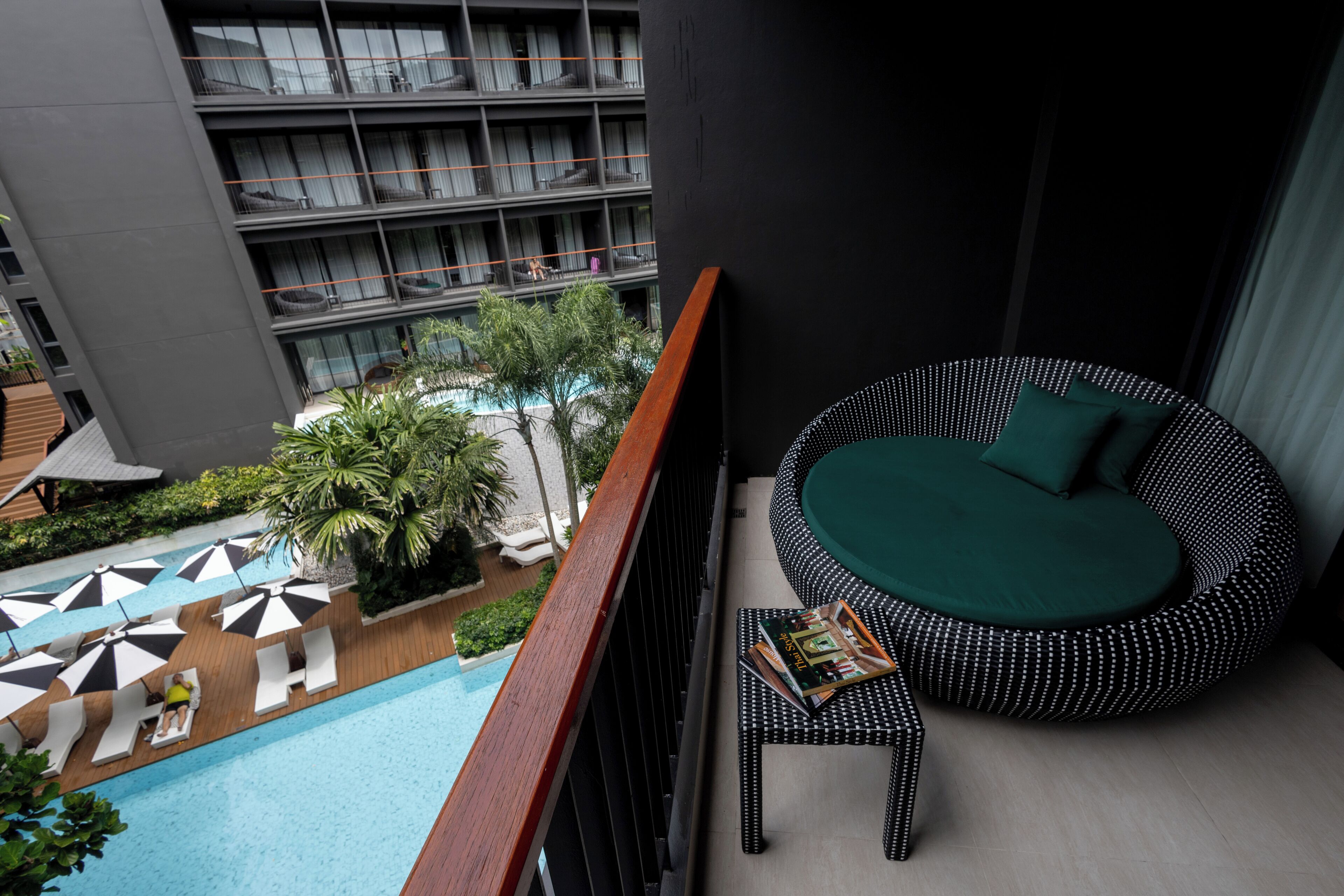 A balcony with a round black and white patterned sofa, a wooden table with a magazine, and a view of a swimming pool and hotel building.