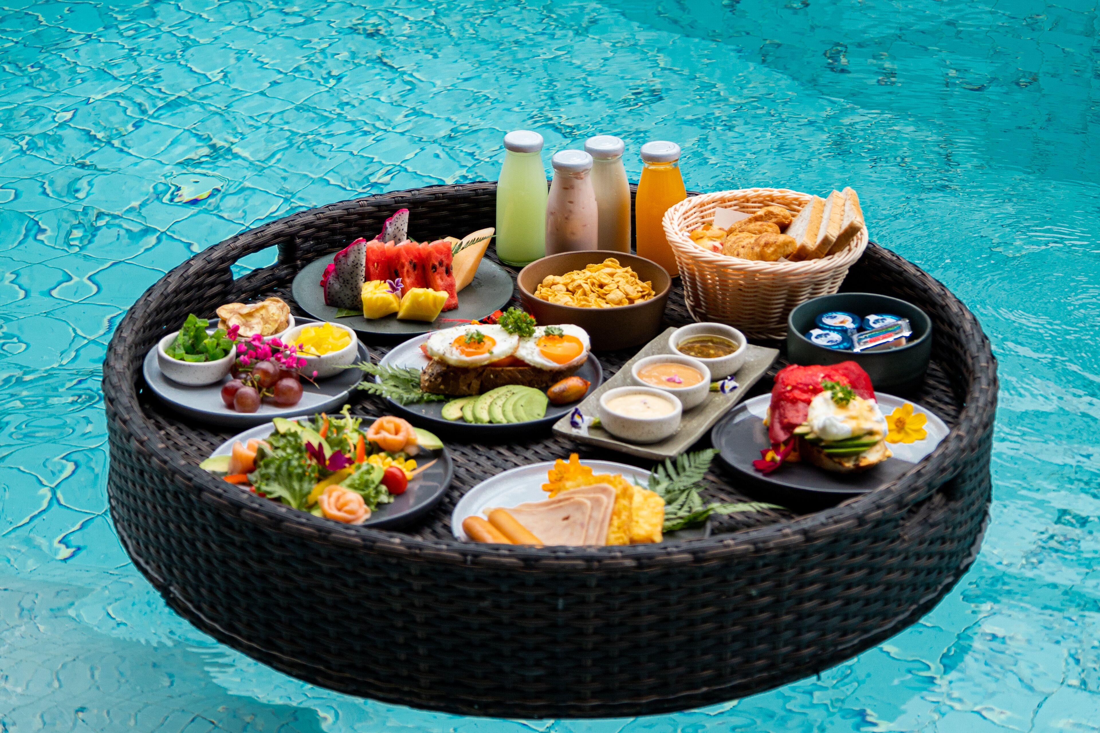 A round wicker tray with various food items and drinks by a pool.