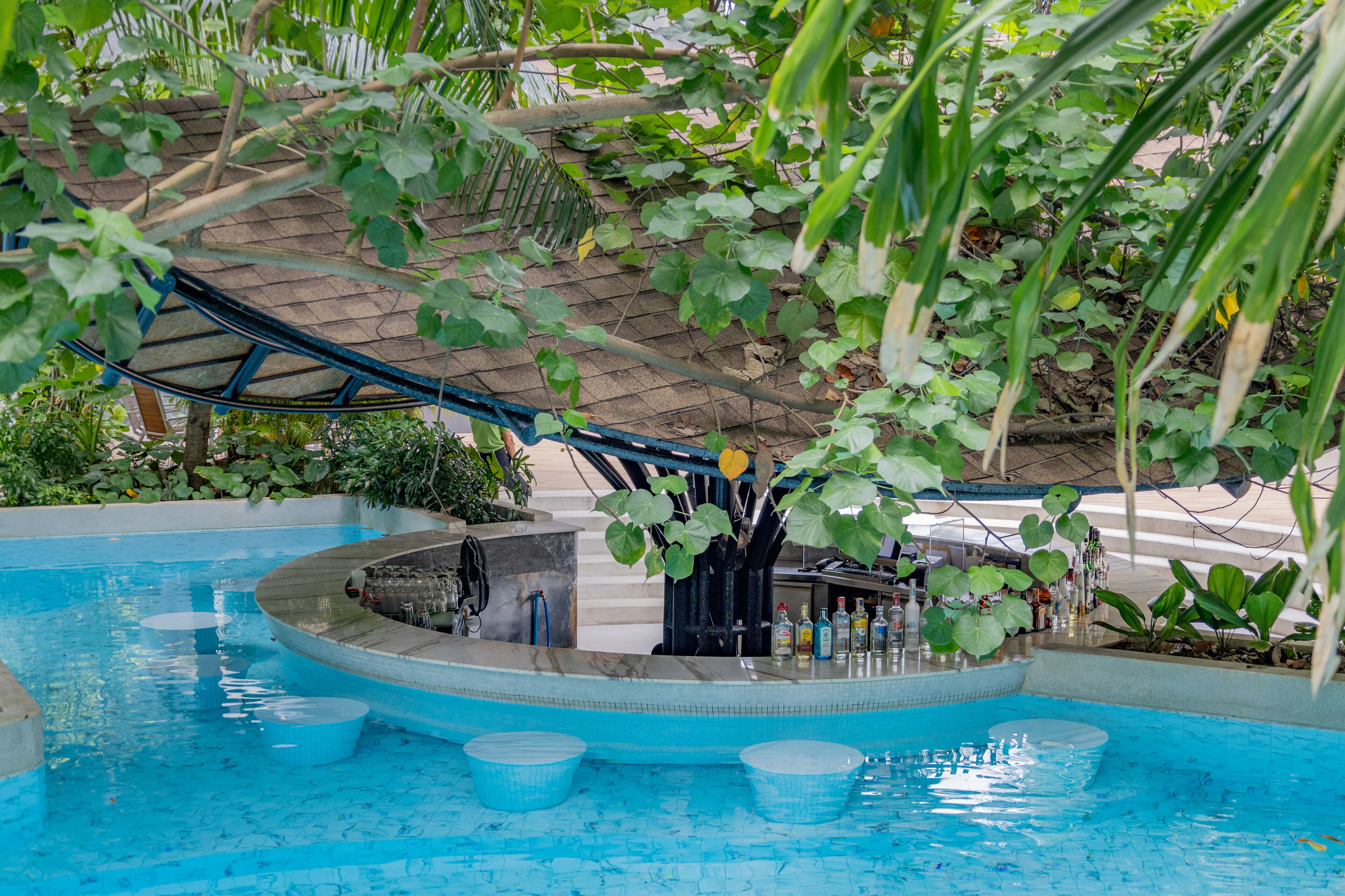 An indoor pool with a bar area, surrounded by lush greenery and a covered seating area.