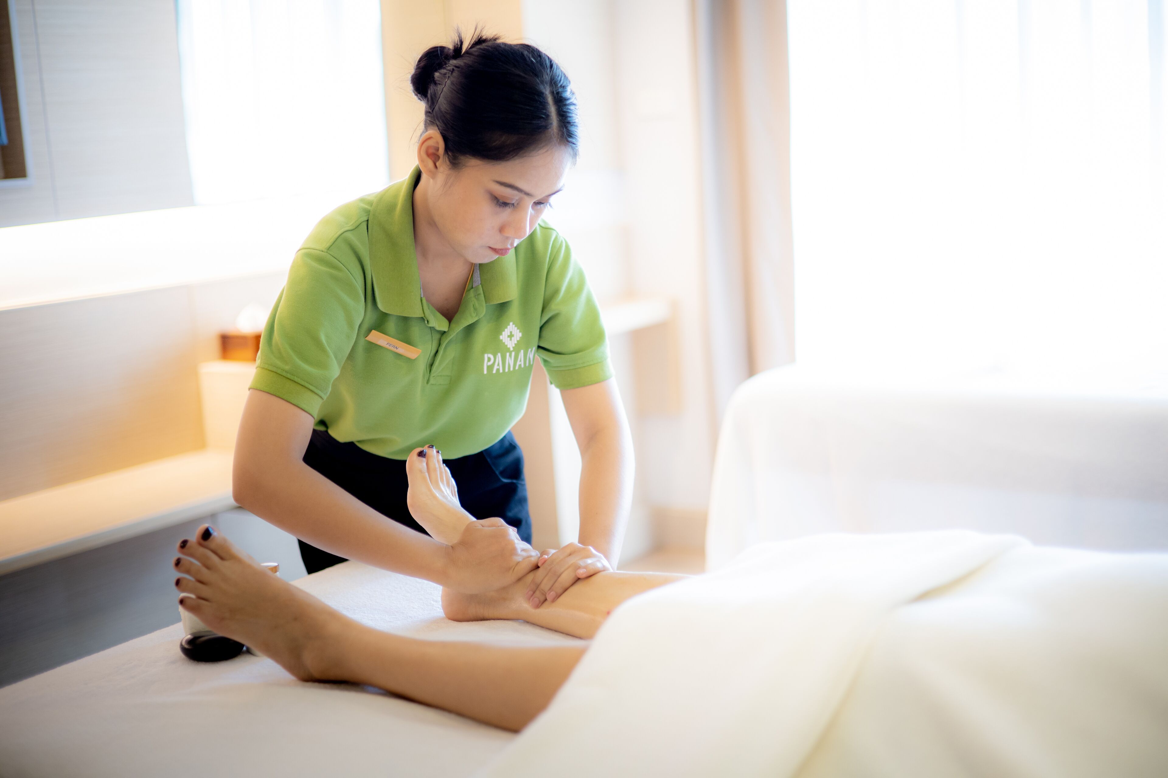 A massage therapist in a green uniform attending to a client on a massage table in a well-lit room with a white bed and a small table with a bottle.